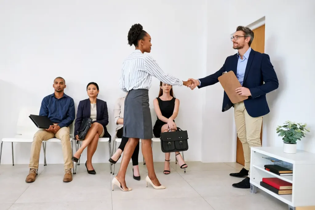 candidata à vaga de emprego aperta a mão de recrutador. Ao fundo, outros candidatos, sentados, observam.